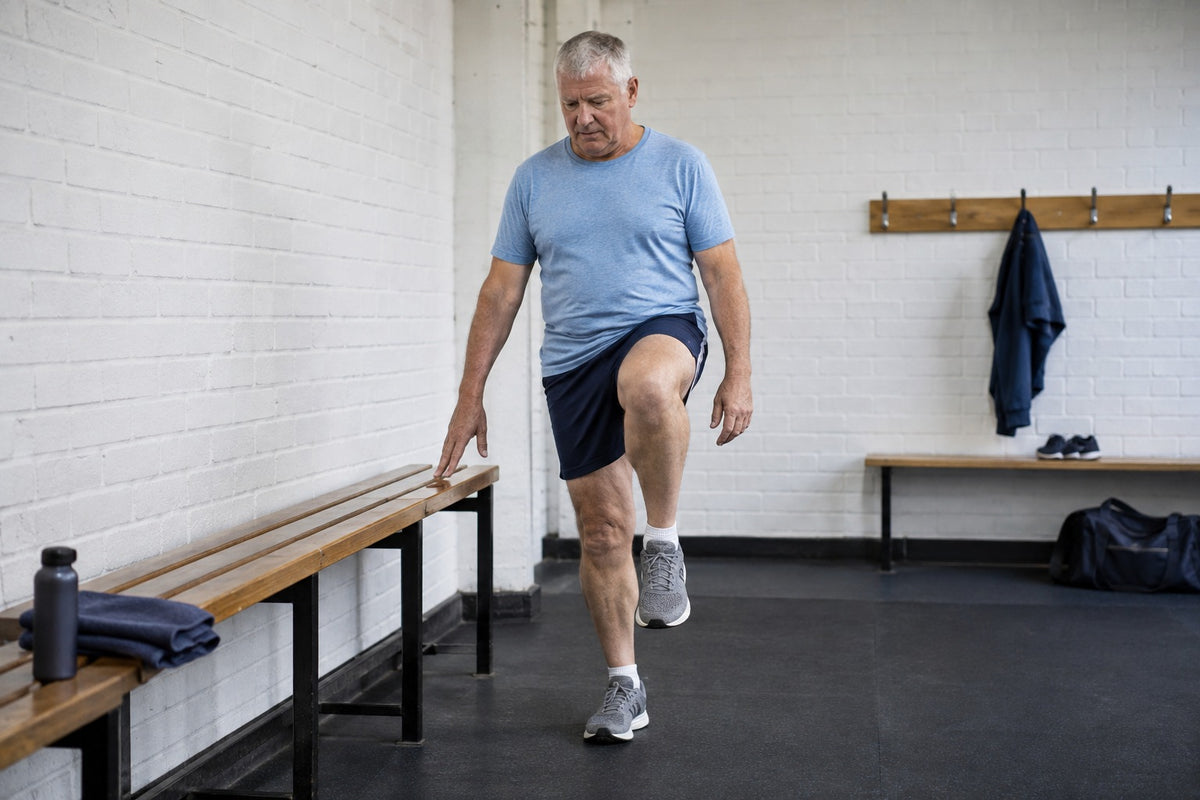 An older man doing a standing knee raise in a changing room, steadying himself with one hand on a bench